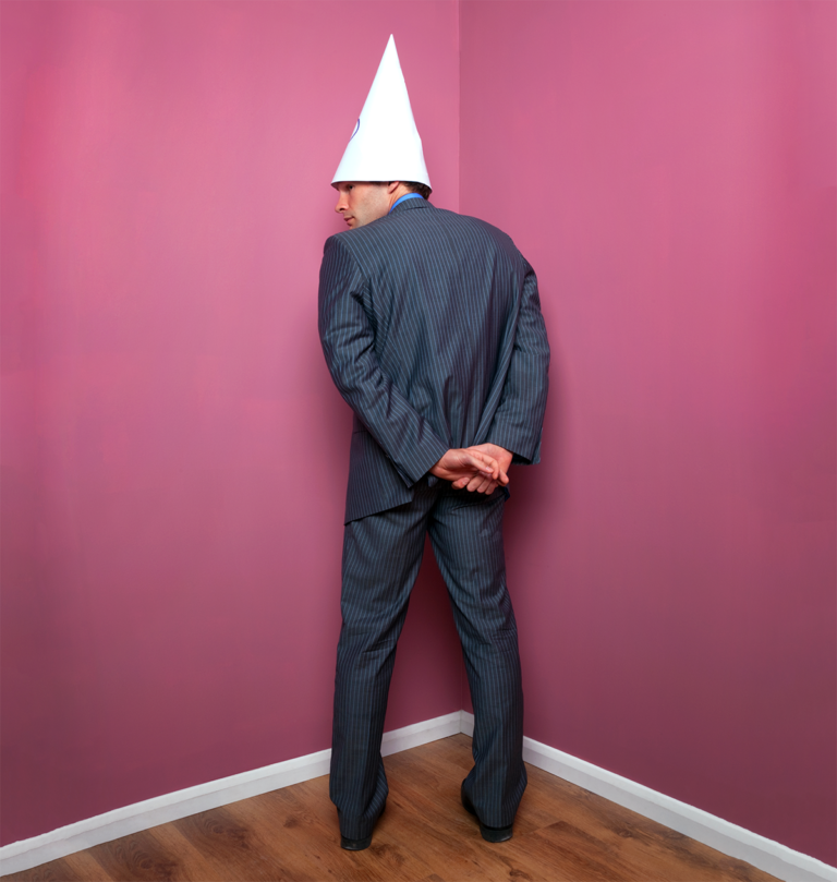 Man standing facing corner of room wearing a dunce cap